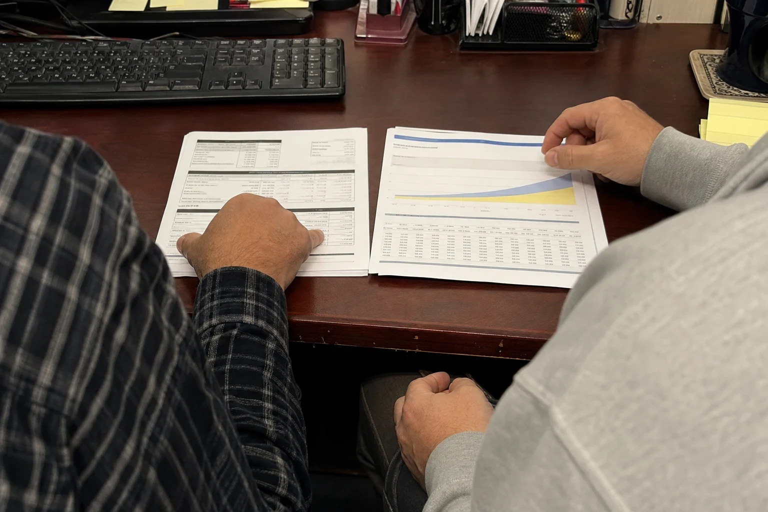 Two people at a wooden office desk comparing a printed statement and a coverage chart with a blue and yellow graph, keyboard and desk organizers behind
